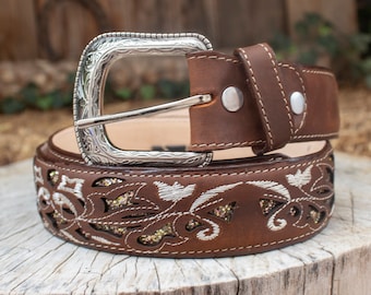 May include: A brown leather belt with a silver buckle and intricate floral embroidery. The embroidery features a white thread and gold glitter accents.
