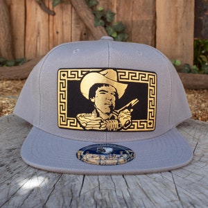 May include: A gray and black baseball cap with a gold-colored patch featuring a man in a cowboy hat holding a gun. The patch is surrounded by a gold-colored Greek key border.