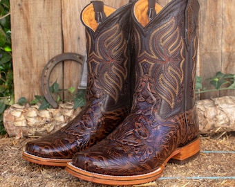 Pode incluir: Um par de botas de cowboy de couro marrom com bordados florais intrincados. As botas têm uma ponta pontiaguda e um salto baixo.