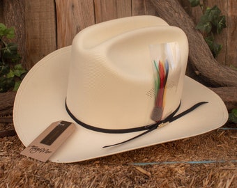 Könnte beinhalten: Ein cremefarbener Strohhut im Cowboy-Stil mit schwarzem Band und einem mehrfarbigen Federschmuck. Der Hut hat ein "Tony Lama"-Etikett am Rand.
