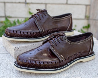 May include: A pair of brown leather shoes with a woven detail around the sole. The shoes have a lace-up closure and a white sole.