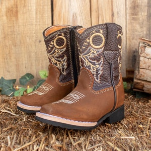 May include: Brown leather children's cowboy boots with a zipper on the side and embroidered floral designs in gold thread.