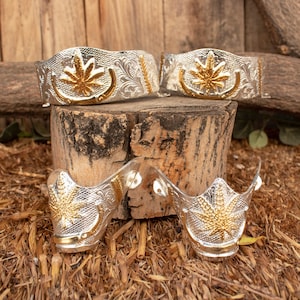 Puede incluir: Dos pares de espuelas de bota de plata y oro con un diseño de hoja. Las espuelas están sobre una superficie de madera.