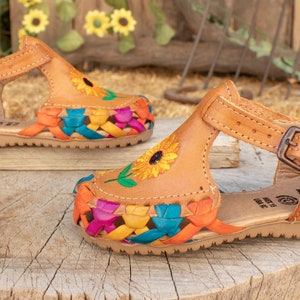 May include: A pair of brown leather sandals with colorful woven straps and embroidered sunflowers. The sandals have an ankle strap with a buckle.