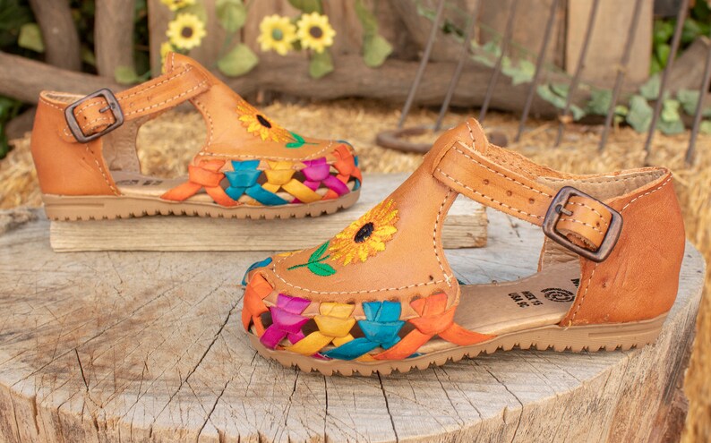 May include: A pair of brown leather sandals with colorful woven straps and embroidered sunflowers. The sandals have an ankle strap with a buckle.