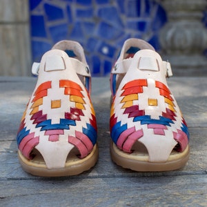 May include: A pair of white leather sandals with a colorful woven pattern. The sandals have a strap that goes around the ankle and a toe loop.