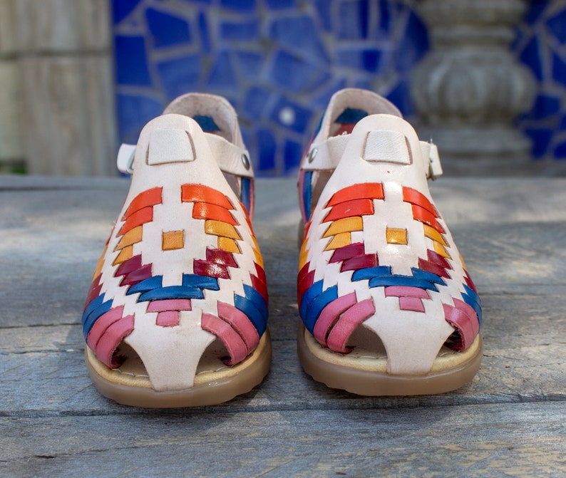 May include: A pair of white leather sandals with a colorful woven pattern. The sandals have a strap that goes around the ankle and a toe loop.