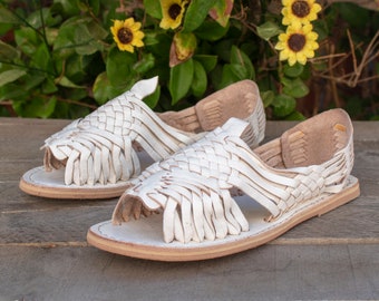 May include: A pair of white leather sandals with a woven design. The sandals have a simple, open-toe design and are made of soft, supple leather.