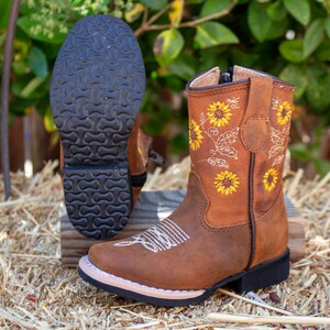 Pu&ograve; includere: Stivali da cowboy in pelle marrone per bambini con cerniera laterale e girasoli ricamati. Gli stivali hanno una suola in gomma nera.