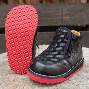 Puede incluir: Botas de tobillo negras de cuero con cremallera lateral y suela de goma roja. Las botas tienen un patrón tejido en la parte superior.