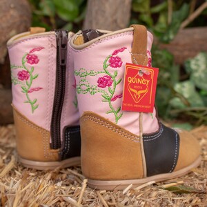 K&ouml;nnte beinhalten: Rosa Leder-Cowboy-Stiefel f&uuml;r Kinder mit Blumenstickerei und braunem Lederabsatz. Die Stiefel haben eine schwarze Lederkappe und einen Rei&szlig;verschluss an der Seite. Die Stiefel haben ein Etikett mit der Aufschrift "Quincy Boots, The Real American Boot".