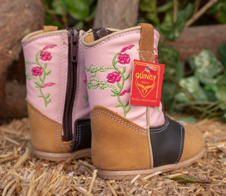K&ouml;nnte beinhalten: Rosa Leder-Cowboy-Stiefel f&uuml;r Kinder mit Blumenstickerei und braunem Lederabsatz. Die Stiefel haben eine schwarze Lederkappe und einen Rei&szlig;verschluss an der Seite. Die Stiefel haben ein Etikett mit der Aufschrift "Quincy Boots, The Real American Boot".