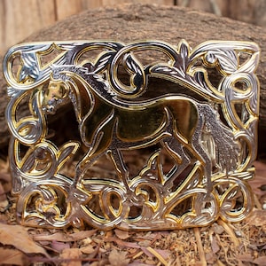 May include: A gold and silver belt buckle with a detailed design of a horse in a floral frame.