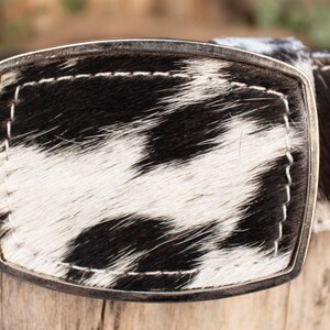 May include: A cowhide belt with a silver buckle. The belt features a black and white spotted pattern, with white stitching along the edges. The buckle is rectangular and has a silver finish.