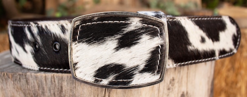 May include: A cowhide belt with a silver buckle. The belt features a black and white spotted pattern, with white stitching along the edges. The buckle is rectangular and has a silver finish.