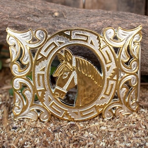 May include: A gold and silver belt buckle with an intricate design featuring a horse's head in the center.