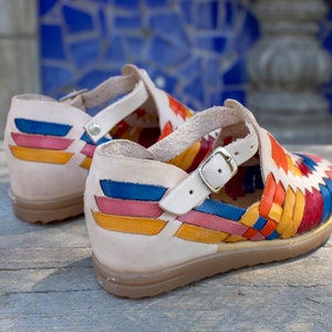 May include: A pair of colorful leather sandals with a woven design. The sandals have a beige leather strap with a buckle and a beige leather sole.
