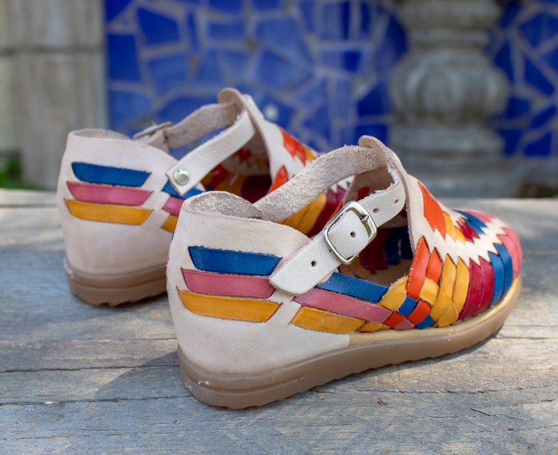 May include: A pair of colorful leather sandals with a woven design. The sandals have a beige leather strap with a buckle and a beige leather sole.