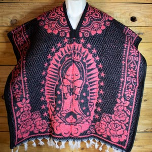 May include: A black and pink poncho with a V-neck and white fringe. The poncho features a central image of a religious figure surrounded by stars and a crown. Floral and geometric patterns adorn the edges.