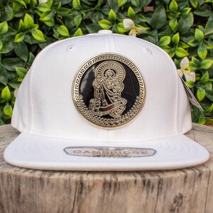 Könnte beinhalten: Eine weiße Baseballkappe mit flachem Schirm. Die Kappe hat ein großes, kreisförmiges Emblem mit einer religiösen Figur in Gold und Schwarz. Im Hintergrund ist grünes Laub zu sehen.