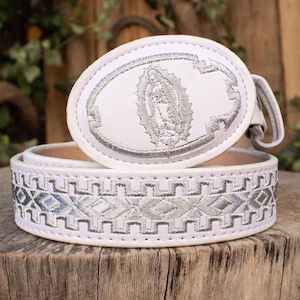 May include: White belt with a silver embroidered geometric pattern and a white oval buckle. The buckle features a silver embroidered image of the Virgin Mary. The belt is resting on a wooden surface.