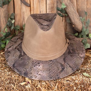 Può includere: Un cappello in stile fedora con un motivo a pelle di serpente sulla tesa e sulla corona e una parte superiore color marrone chiaro. Il cappello ha una fascia in pelle attorno alla base della corona. Il cappello è appoggiato su un letto di fieno.