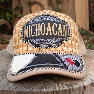 May include: A tan and brown trucker hat with a woven pattern. The hat features a black patch with the word "MICHOACAN" in gold lettering. The brim has a black and white design with a red and black graphic.