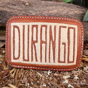 May include: Rectangular leather belt buckle with the word "DURANGO" in large, bold, brown letters. The letters are on a cream-colored background, framed by a brown leather border with white stitching. The buckle is a rustic accessory.