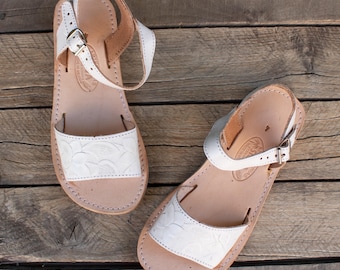 May include: Two white leather sandals with ankle straps and buckle closures. The sandals have a natural leather footbed and a decorative embossed design on the toe strap. The sandals are displayed on a wooden surface.