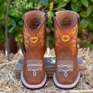 Pu&ograve; includere: Stivali in pelle marrone per bambini con una cerniera laterale. Gli stivali hanno un ricamo a forma di girasole e il testo "GENUINE LEATHER MADE IN MEXICO" all'interno.