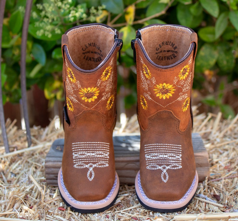 Pu&ograve; includere: Stivali in pelle marrone per bambini con una cerniera laterale. Gli stivali hanno un ricamo a forma di girasole e il testo "GENUINE LEATHER MADE IN MEXICO" all'interno.