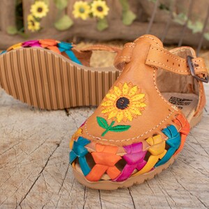 May include: A pair of brown leather sandals with colorful woven straps and an embroidered sunflower design. The sandals have an ankle strap with a buckle closure.