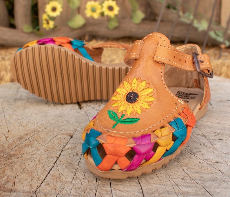 May include: A pair of brown leather sandals with colorful woven straps and an embroidered sunflower design. The sandals have an ankle strap with a buckle closure.