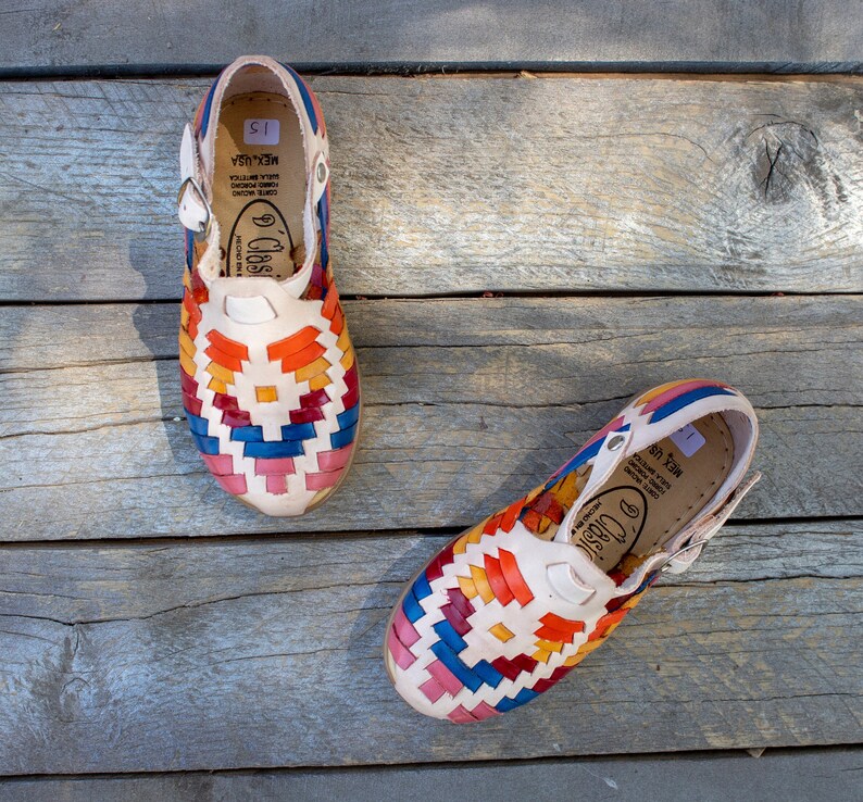 May include: A pair of white leather sandals with colorful woven straps. The sandals have a buckle closure and the text "Clasico" and "Mex. USA" is printed on the straps.