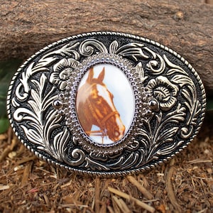 May include: Oval-shaped silver-tone belt buckle with a detailed floral and scroll design. The center features an oval image of a brown horse's head. The buckle has a black border and is set against a natural wood background.