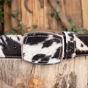 May include: A black and white cowhide belt with a silver buckle. The belt features a rectangular buckle and is made of genuine cowhide. The black and white pattern is consistent throughout the belt, and the stitching is white.
