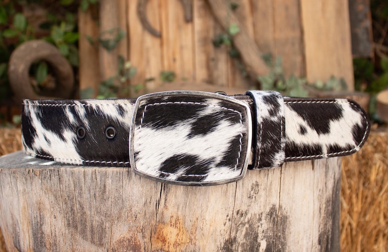 May include: A black and white cowhide belt with a silver buckle. The belt features a rectangular buckle and is made of genuine cowhide. The black and white pattern is consistent throughout the belt, and the stitching is white.