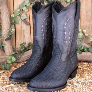 Pode incluir: Um par de botas de cowboy pretas com costuras intrincadas e um padrão texturizado no pé e na haste. As botas têm bico fino e salto empilhado, contra um fundo de madeira rústica.