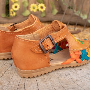 May include: A pair of brown leather sandals with colorful woven straps and a buckle closure. The sandals have a floral embroidery design.