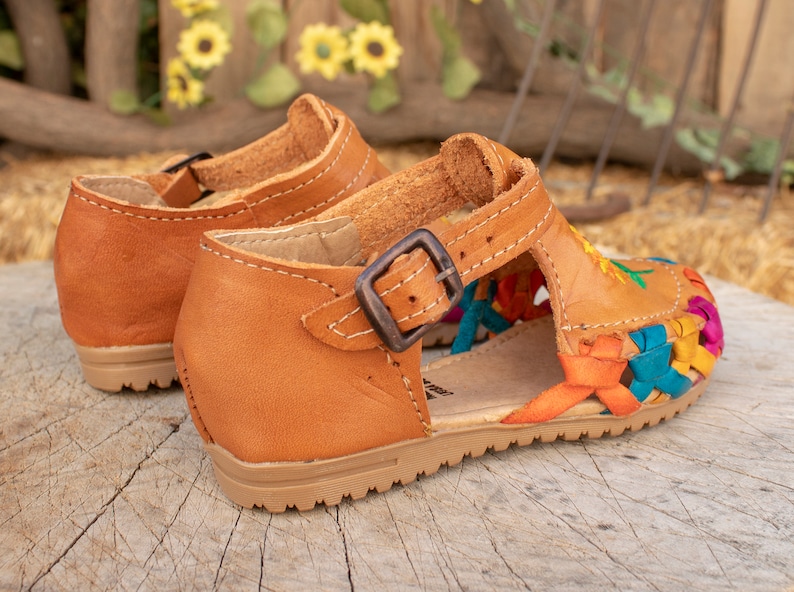 May include: A pair of brown leather sandals with colorful woven straps and a buckle closure. The sandals have a floral embroidery design.