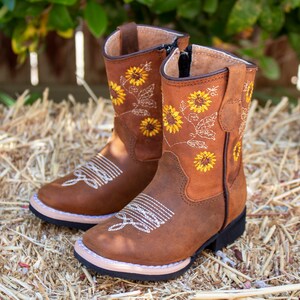 Pu&ograve; includere: Stivali da cowboy in pelle marrone con ricamo a girasole. Gli stivali hanno una cerniera laterale e una punta a punta.