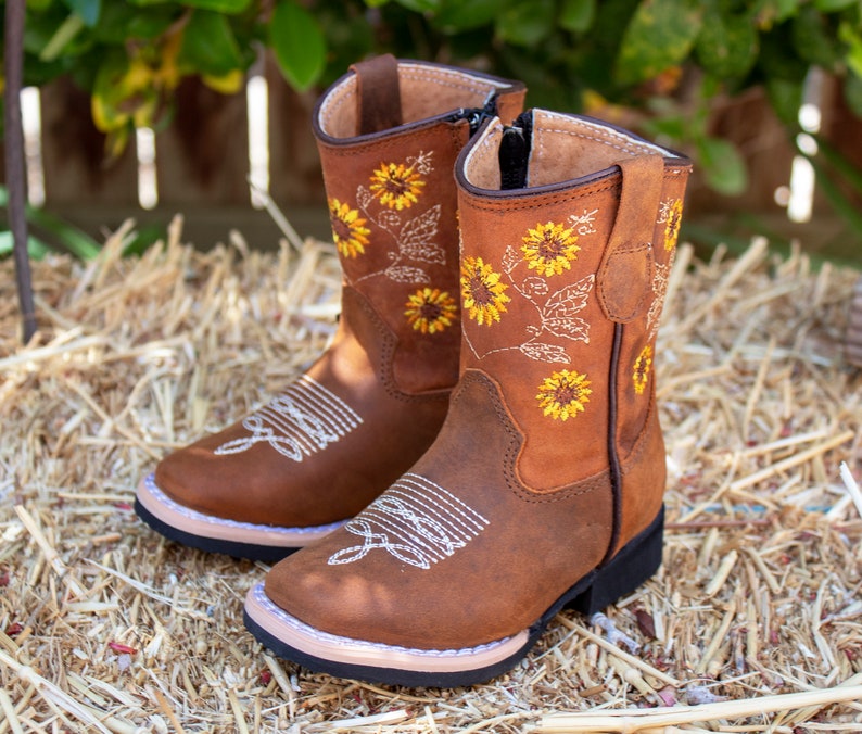 Pu&ograve; includere: Stivali da cowboy in pelle marrone con ricamo a girasole. Gli stivali hanno una cerniera laterale e una punta a punta.