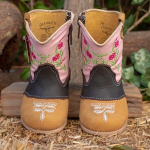 K&ouml;nnte beinhalten: Ein Paar rosa und braune Cowboystiefel mit Blumenstickerei und Libellenmotiven. Die Stiefel haben einen Rei&szlig;verschluss an der Seite und einen braunen Lederschaft.