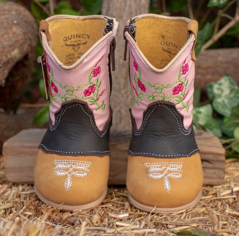 K&ouml;nnte beinhalten: Ein Paar rosa und braune Cowboystiefel mit Blumenstickerei und Libellenmotiven. Die Stiefel haben einen Rei&szlig;verschluss an der Seite und einen braunen Lederschaft.