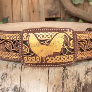 May include: A brown leather belt with a gold-colored rooster buckle. The belt is decorated with a Celtic knot design and leaves.