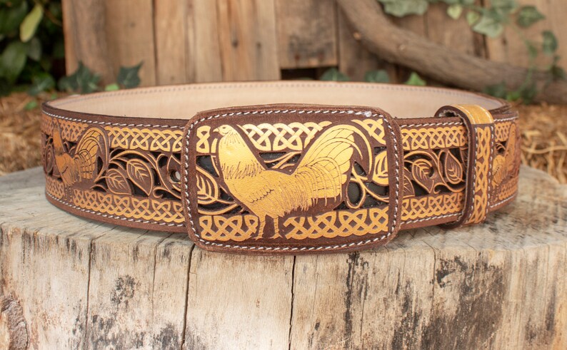 May include: A brown leather belt with a gold-colored rooster buckle. The belt is decorated with a Celtic knot design and leaves.