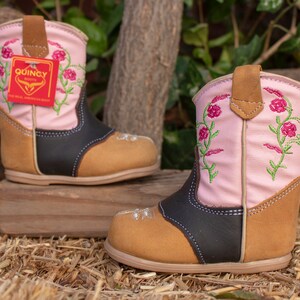K&ouml;nnte beinhalten: Ein Paar rosa und braune Cowboystiefel mit Blumenstickerei. Die Stiefel haben einen schwarzen Lederbesatz und eine braune Lederlasche. Die Stiefel stehen auf einem Strohboden.