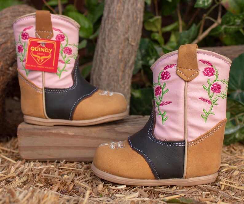 K&ouml;nnte beinhalten: Ein Paar rosa und braune Cowboystiefel mit Blumenstickerei. Die Stiefel haben einen schwarzen Lederbesatz und eine braune Lederlasche. Die Stiefel stehen auf einem Strohboden.