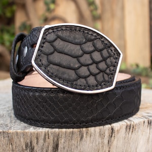 May include: A black leather belt with a silver buckle. The belt features a textured, scale-like pattern. The rectangular buckle has a silver border and a matching scale-like texture. The belt is displayed on a wooden surface.