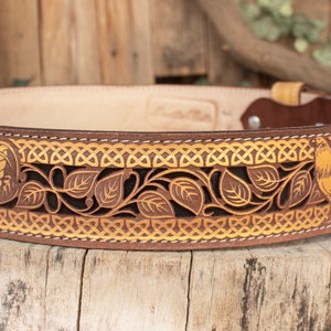 May include: A brown leather belt with a gold-colored Celtic knot design and two roosters. The belt is decorated with intricate cutouts of leaves and vines.
